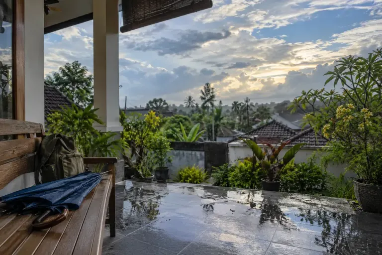 Teras rumah dengan payung, tanaman tropis dan langit berawan setelah hujan.